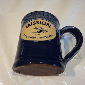 May include: A dark blue ceramic mug with a large handle. The mug features a tan oval with the words "MISSION SAN JUAN CAPISTRANO 1776" and a swallow illustration. The rim has a darker blue speckled design.