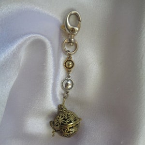 May include: A silver-toned metal lobster clasp keychain featuring a vertical chain with a small gold-toned circular bead, a silver-toned circular bead with a clear center, and a round, hollow, gold-toned metal cage pendant with an intricate cutout pattern.