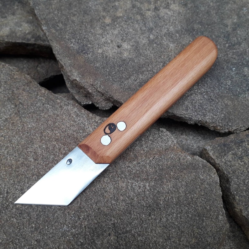 May include: A small, silver, utility knife with a wooden handle. The handle has a dark brown stain and a circular design with a white circle in the center.