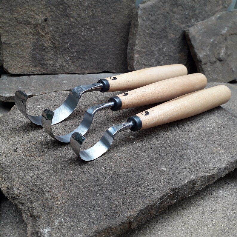 May include: Three wood carving tools with curved blades and wooden handles. The tools are arranged in a stack, with the largest tool on the bottom and the smallest tool on top. The tools are made of metal and wood and have a natural finish.