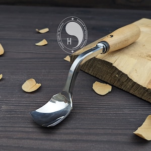 May include: A curved wood carving tool with a metal blade and a wooden handle. The tool is resting on a piece of wood with wood shavings around it.