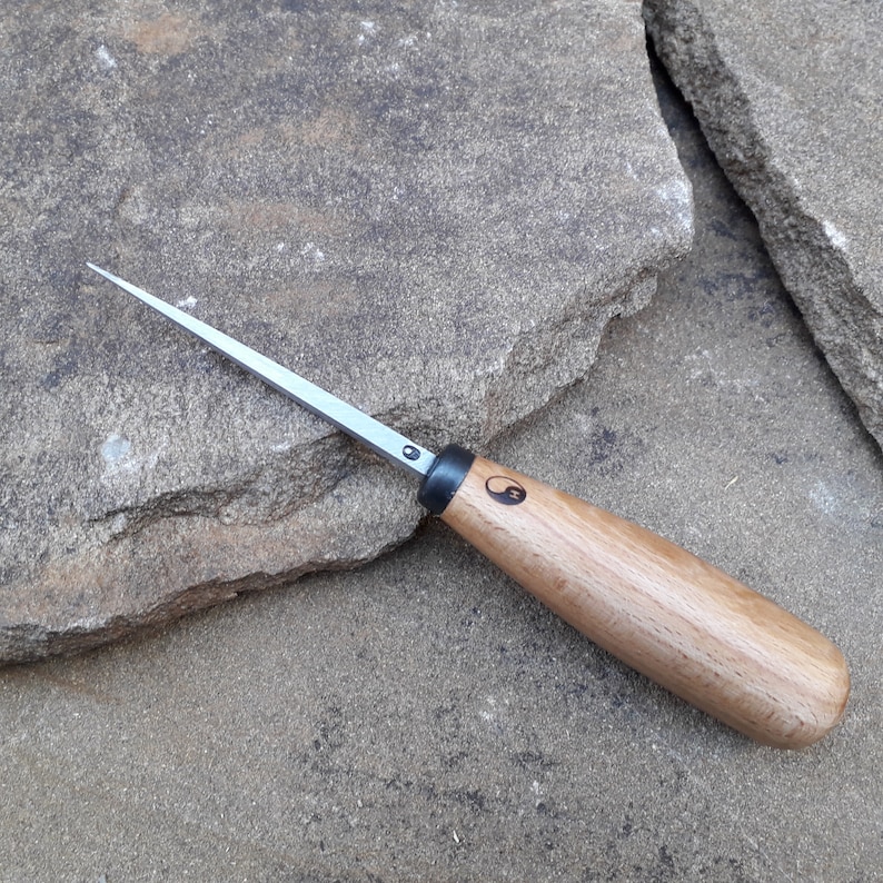 May include: A close-up of a wood carving tool with a thin, sharp blade and a wooden handle. The handle is marked with a yin-yang symbol.