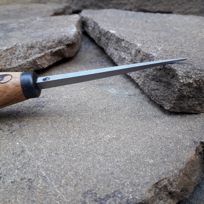 May include: A close-up of a wood-handled carving tool with a sharp, pointed blade. The tool is resting on a rough, gray stone surface.