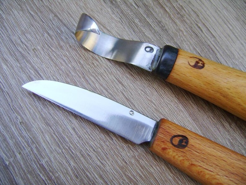 Op de afbeelding: Twee snijmesjes met houten handvatten en zilveren messen. Het mes aan de linkerkant heeft een gebogen mes en het mes aan de rechterkant heeft een recht mes.