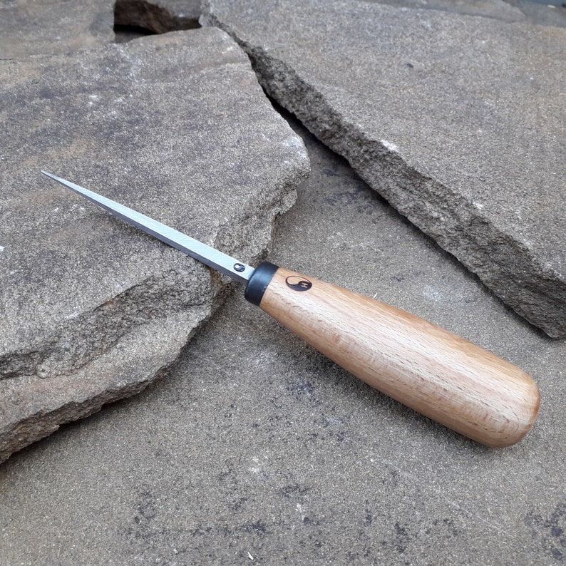 May include: A small, wooden-handled carving tool with a sharp, pointed blade. The handle is made of light-colored wood and has a black yin-yang symbol engraved on it.