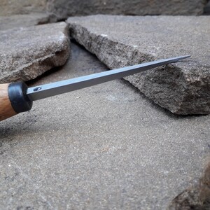 May include: A close-up of a wood and metal carving tool with a sharp, pointed blade. The tool is resting on a rough, gray stone surface.