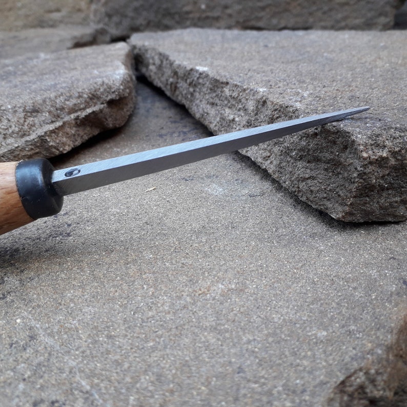 May include: A close-up of a wood and metal carving tool with a sharp, pointed blade. The tool is resting on a rough, gray stone surface.
