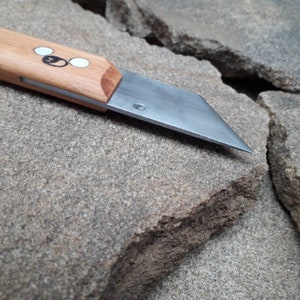 May include: A close-up of a small, wooden-handled knife with a silver blade. The blade is sharp and pointed, and the handle is made of light brown wood. The knife is resting on a rough, gray stone surface.