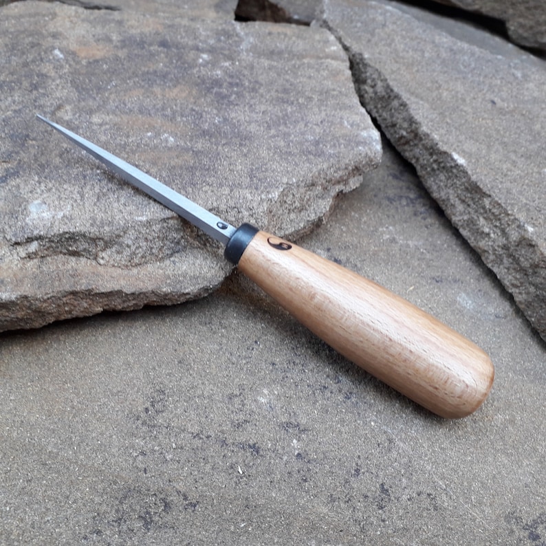 May include: A small, wooden-handled carving tool with a sharp, pointed blade. The tool is made of metal and wood and has a black handle with a small, engraved symbol.