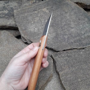May include: A close-up of a wooden-handled knife with a black blade. The knife is held in a hand, and the blade is pointed towards the top of the image.
