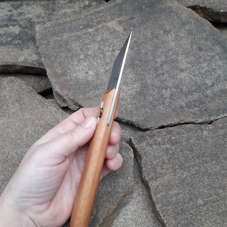 May include: A close-up of a wooden-handled knife with a black blade. The knife is held in a hand, and the blade is pointed towards the top of the image.