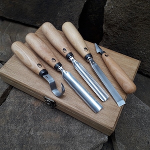 May include: Set of six wood carving tools with wooden handles and metal blades. The tools are arranged in a wooden box.