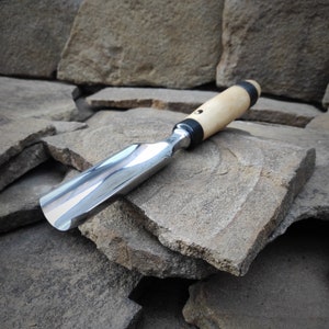 May include: A close-up of a wood and metal chisel with a black and white handle. The chisel is resting on a pile of gray rocks.