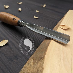 May include: A wood carving gouge with a curved metal blade and a smooth, light brown wooden handle. The handle features a small circular logo with the letter H. The tool is positioned near a piece of light-colored wood with scattered shavings.