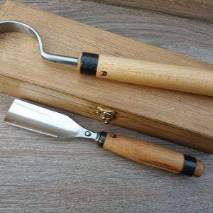 Może przedstawiać: Dwa narzędzia do rzeźbienia w drewnie z metalowymi ostrzami i drewnianymi uchwytami spoczywają na drewnianej skrzynce. Jedno narzędzie ma zakrzywione ostrze, drugie płaskie. Skrzynka jest prostokątna z mosiężnym zapięciem.