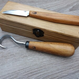 Op de afbeelding: Twee houtsnijgereedschappen met houten handvatten in een houten doos. De gereedschappen zijn een klein mes en een haakmes.