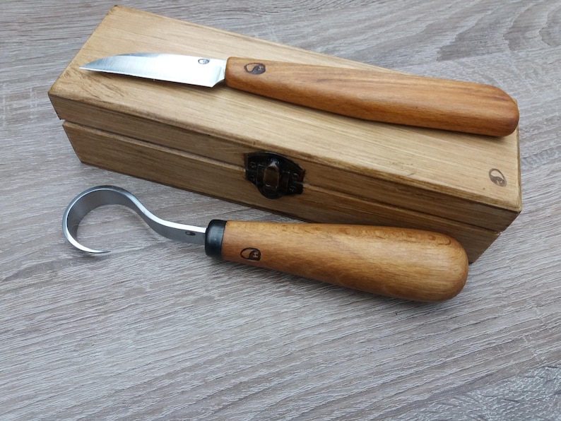 Op de afbeelding: Twee houtsnijgereedschappen met houten handvatten in een houten doos. De gereedschappen zijn een klein mes en een haakmes.