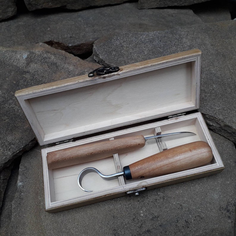 Op de afbeelding: Twee houtsnijgereedschappen met houten handvatten in een houten doos met een sluiting. De gereedschappen zijn een haakmes en een recht mes.