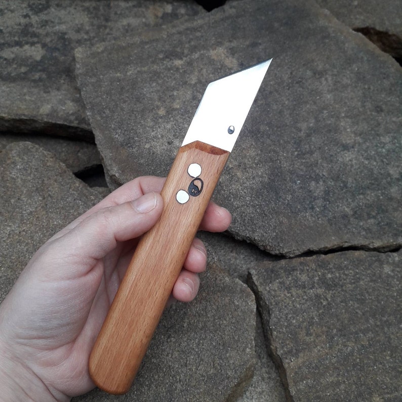 May include: A wooden handled utility knife with a silver blade. The handle has two silver rivets and a small silver circle with a design on it.