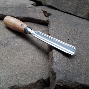 May include: A close-up of a wood handled carving tool with a silver blade. The tool is lying on a rough, gray stone surface.