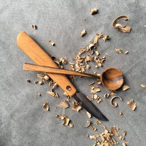 Op de afbeelding: Een houten lepel en een houten handvatmes met een metalen mes, beide liggend op een grijs oppervlak met houtkrullen eromheen.