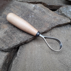 May include: A wooden handled carving tool with a metal loop for shaping wood. The tool is lying on a rough, gray stone surface.