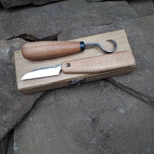 Op de afbeelding: Twee houtsnijgereedschappen met houten handvatten in een houten doos. De gereedschappen zijn een klein mes en een haakmes.