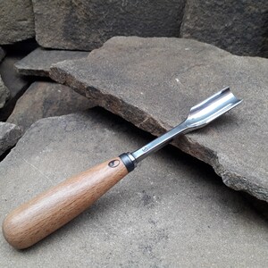 以下が含まれることがあります： 湾曲した研磨された鋼の刃と明るい茶色の木製ハンドルを備えた木彫りのガウジ。 このツールは木材を成形するように設計されており、刃がハンドルに接する場所に金属製のフェルールが付いています。 ガウジは灰色の石の背景にあります。