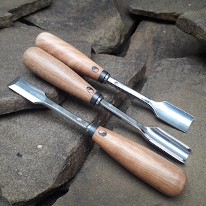 Könnte beinhalten: Drei Holzschnitzwerkzeuge mit einem Yin-Yang-Symbol auf den Griffen. Die Werkzeuge haben unterschiedliche Klingenformen und -größen. Die Werkzeuge liegen auf einer Oberfläche aus rauen, grauen Steinen.