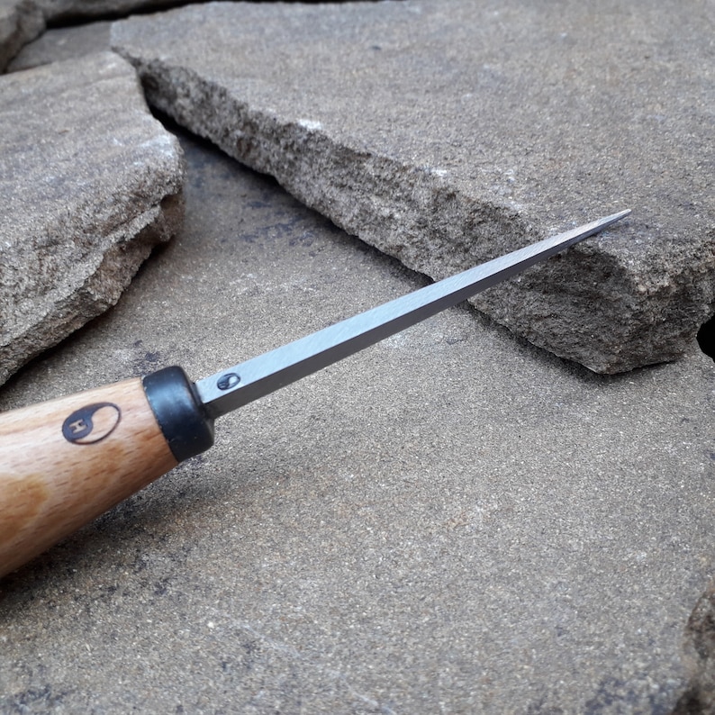 May include: A close-up of a wood-handled carving tool with a thin, sharp blade. The tool has a black handle with a yin-yang symbol and the letter "H" engraved on it.