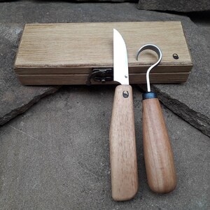 Op de afbeelding: Twee houtsnijgereedschappen met houten handvatten in een houten doos. De gereedschappen hebben een metalen mes en een haak.