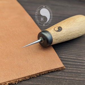 May include: A close-up of a leather working tool with a wooden handle and a sharp metal point. The tool is resting on a piece of brown leather. The handle is engraved with a yin-yang symbol and the text "Forged by Humanity".