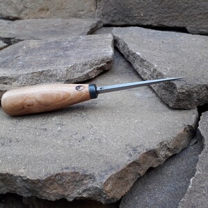 May include: A close-up of a wood handled carving tool with a thin, sharp blade. The tool is resting on a pile of gray rocks.