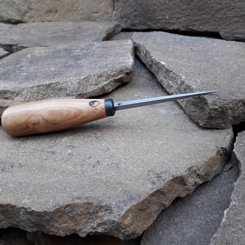 May include: A close-up of a wood handled carving tool with a thin, sharp blade. The tool is resting on a pile of gray rocks.