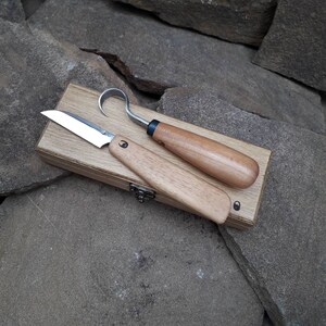 Op de afbeelding: Twee houtsnijgereedschappen met houten handvatten in een houten doos. De gereedschappen zijn een klein mes en een haakmes.