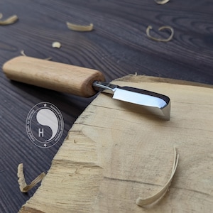 Peut inclure: Un couteau à sculpter le bois avec une lame en métal poli et un manche en bois clair. L'outil est posé sur une pièce de bois clair avec des copeaux visibles à proximité. Un logo circulaire avec la lettre H est visible sur la lame.