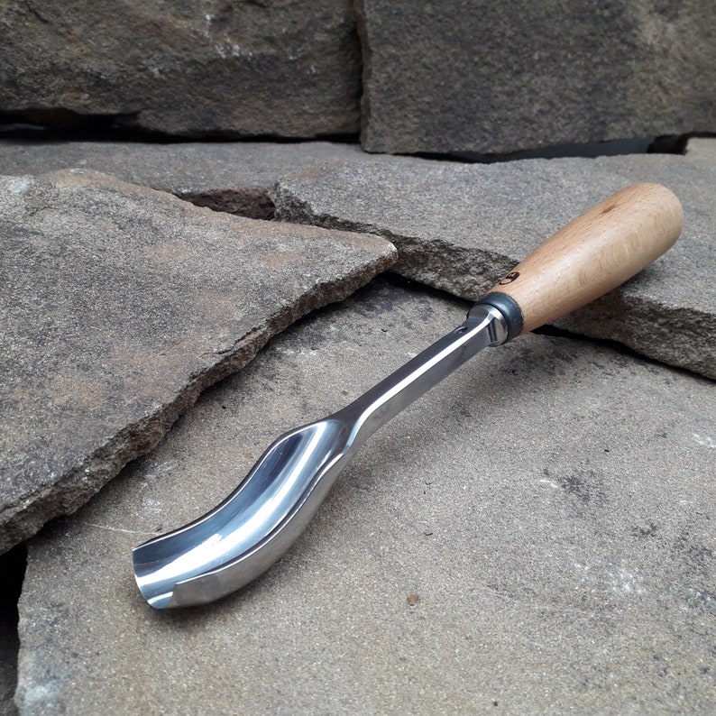 以下が含まれることがあります： 木製のハンドルと金属製の刃を持つ湾曲した木彫りツール。ツールは灰色の石の積み重ねの上に置かれています。