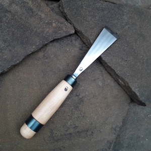 May include: A wood carving chisel with a light-colored wooden handle and a dark metal band. The blade is made of shiny metal. The chisel is lying on a dark, textured stone surface.