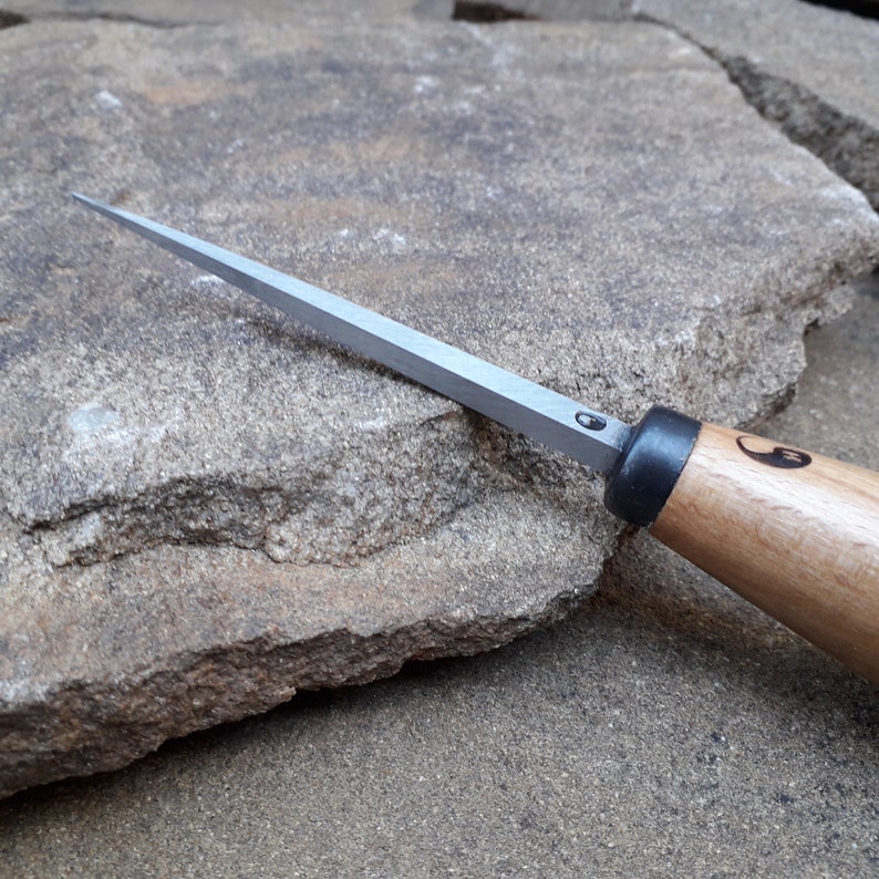 May include: A close-up of a wood carving knife with a silver blade and a wooden handle. The handle has a black ring and a yin-yang symbol.