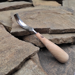 May include: A curved wood carving gouge with a polished steel blade. The blade has a curved shape and a patterned design. The handle is made of light-colored wood and features a small circular emblem. The tool is designed for woodworking and carving.