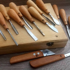 May include: A selection of wood carving tools with wooden handles and stainless steel blades. The tools are arranged on and around a wooden box. Two knives with angled blades are in the foreground. The handles have a circular logo.
