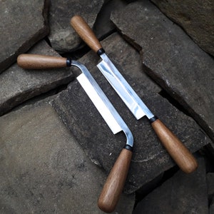 May include: Two wooden handled metal woodworking tools, a scraper and a plane, are lying on a pile of gray and brown rocks.