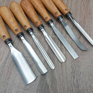May include: A set of six wood carving chisels with polished metal blades and light brown wooden handles. Each handle features a small circular logo stamped near the metal ferrule. The tools vary in blade width and shape, including flat and gouge tips.