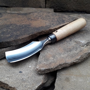 May include: A curved wood carving knife with a wooden handle and a metal blade. The blade is curved and has a sharp edge. The handle is made of wood and has a smooth finish. The knife is resting on a pile of rocks.