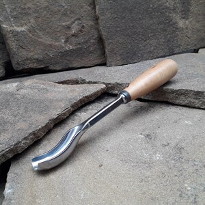 以下が含まれることがあります： 木製のハンドルと金属製の刃を持つ湾曲した木彫りツール。ツールは灰色の石の表面に置かれています。