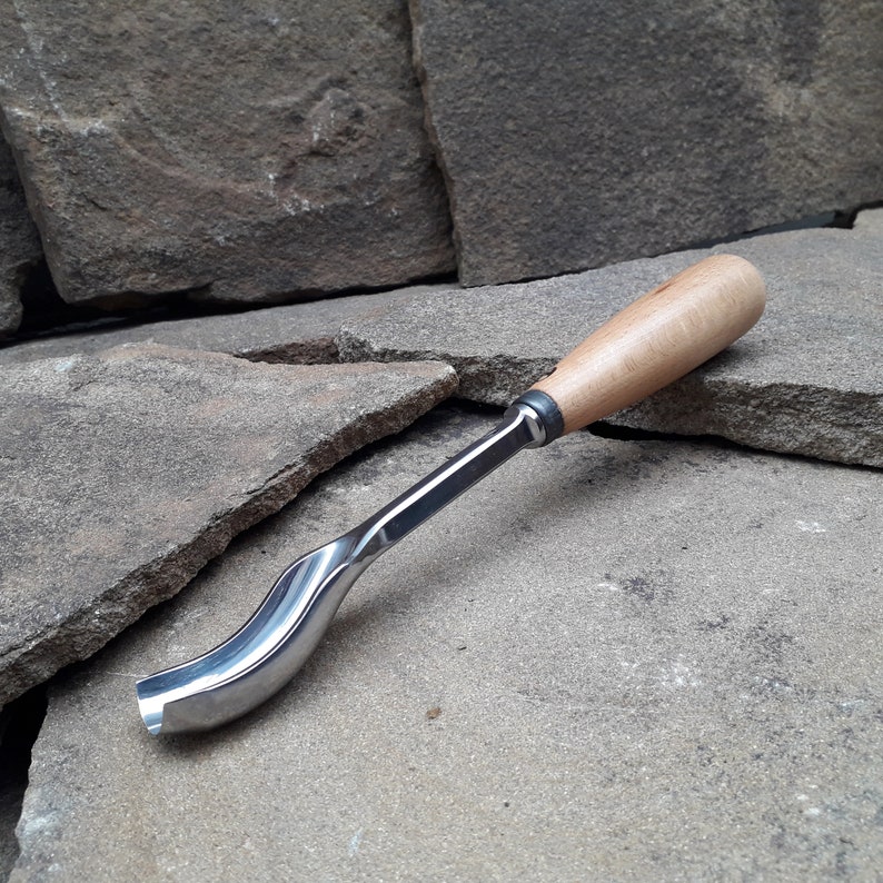 以下が含まれることがあります： 木製のハンドルと金属製の刃を持つ湾曲した木彫りツール。ツールは灰色の石の表面に置かれています。