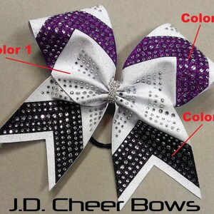 May include: A white, purple, and black cheer bow with rhinestones. The bow has a chevron pattern and is tied with a black elastic band.
