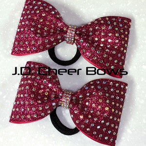 May include: Two burgundy cheer bows with rhinestones and black hair elastics. The bows are made of sparkly fabric and have a satin finish.