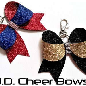 May include: Two glittery cheer bows with silver keychains. One bow is red and blue, the other is black and gold. Both bows have a rhinestone band across the center. The text "J.D. Cheer Bows" is at the bottom.