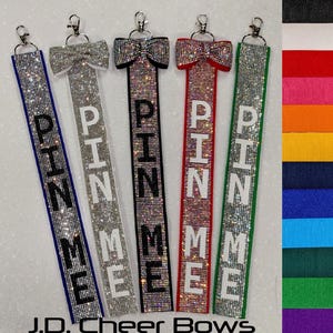 May include: Five cheer bow straps with rhinestones and the word "PIN ME" on each strap. The straps are in different colors: black, white, iridescent, red, and green. The straps are hanging from a white background. J.D. Cheer Bows is written at the bottom of the image.